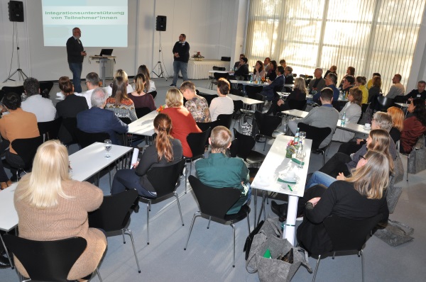 Viele Menschen sitzen in einem Raum und schauen auf zwei vorne stehende Männer, hinter denen eine Bildschirmpräsentation zu sehen ist.