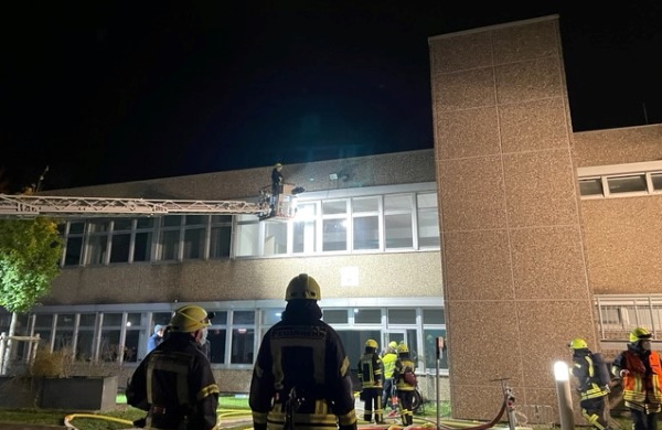 Ein großes Gebäude bei Nacht, vor dem einige Feuerwehrleute mit Feuerwehrschlauch, etc. stehen und eine Feuerwehrleiter, die in den ersten Stock ausgefahren ist.