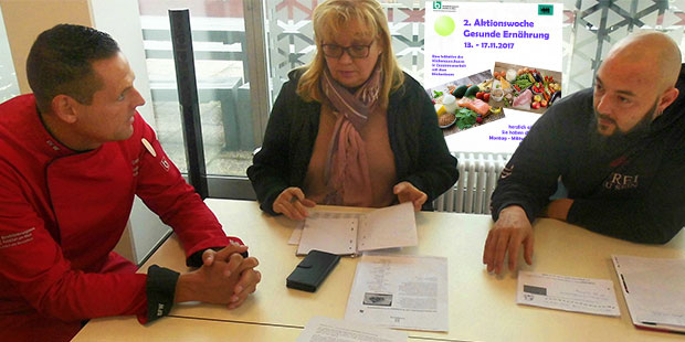 Drei Personen sitzen an einem Tisch, auf dem verschiedene Ausdrucke liegen und unterhalten sich.