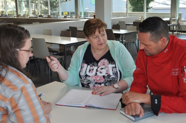 Drei Personen sitzen in einem großen Raum mit mehreren Tischen mit Notizblöcken an einem Tisch und unterhalten sich.