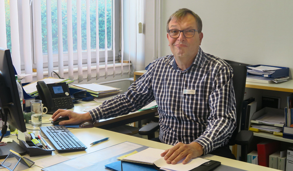 Mittelalter Mann mit Brille sitzt an einem vollen Schreibtisch und schaut in die Kamera.