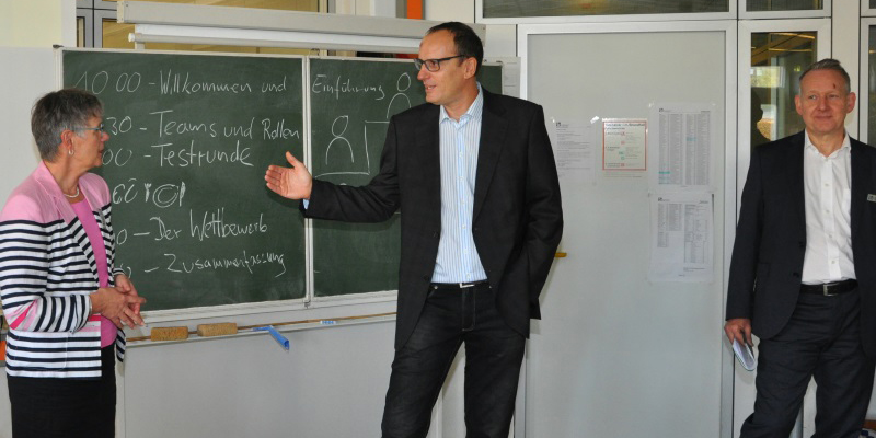 Drei Personen stehen in einem Klassenzimmer vor einer Tafel und unterhalten sich.