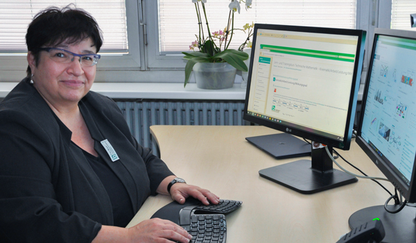 Eine mittelalte Frau sitzt an einem Schreibtisch vor zwei Monitoren und schaut in die Kamera.