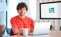 Ein junger Mann sitzt lächelnd an einem Schreibtisch mit Laptop und Tasse. Ein Bildschirm im Hintergrund zeigt das Logo des BFW-Frankfurt.