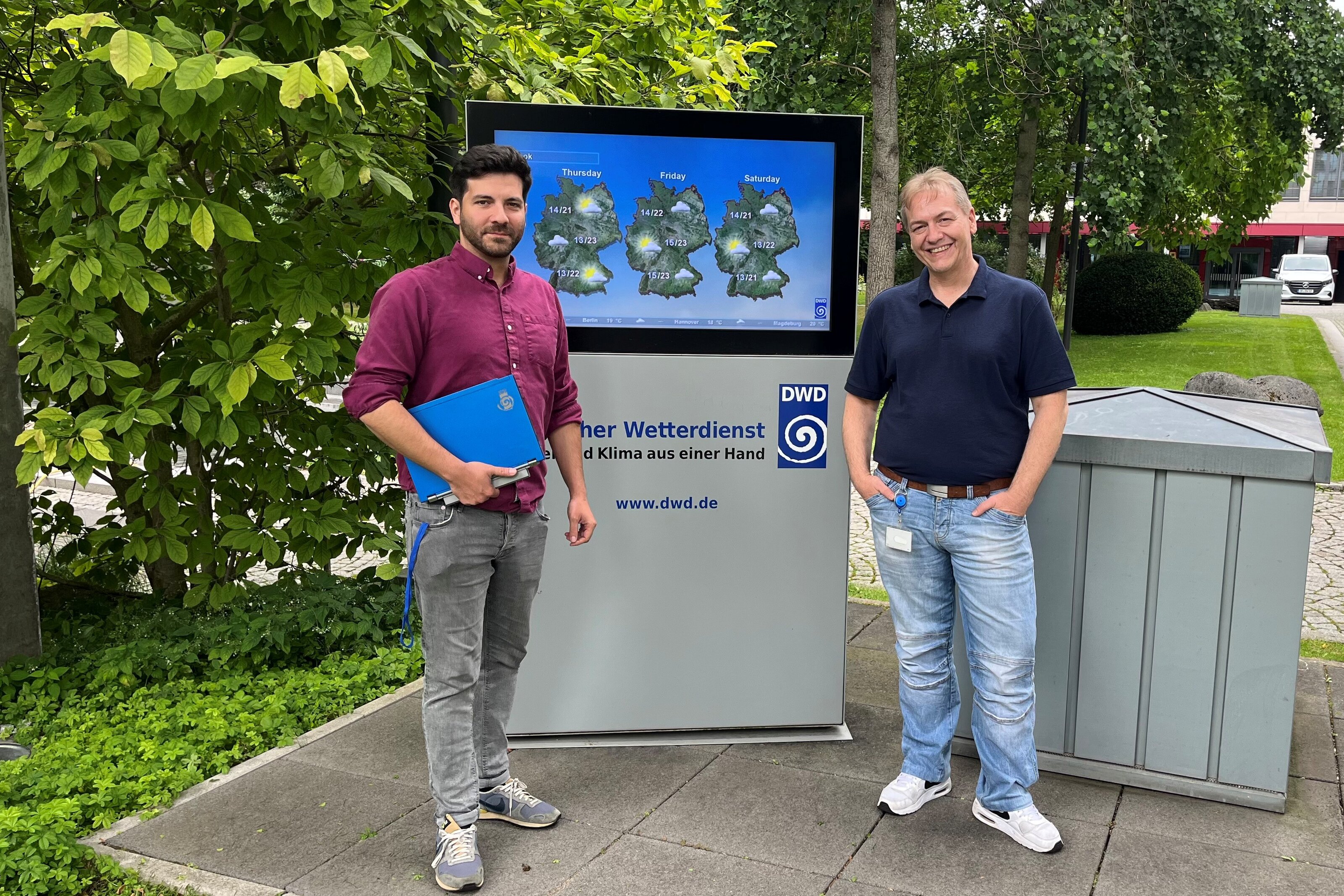 Sascha und Stefan stehen vor dem Gebäude des Deutschen Wetterdienstes, direkt hinter ihnen ein Monitor mit den aktuellen Wetterdaten.