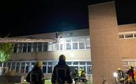 Ein großes Gebäude bei Nacht, vor dem einige Feuerwehrleute mit Feuerwehrschlauch, etc. stehen und eine Feuerwehrleiter, die in den ersten Stock ausgefahren ist.